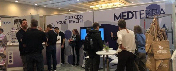 Trade show booth with people interacting. Booth displays text 'OUR CBD. YOUR HEALTH.' and 'MEDTERRA'. Tote bags with 'MEDTERRA' logo hang on the right side.