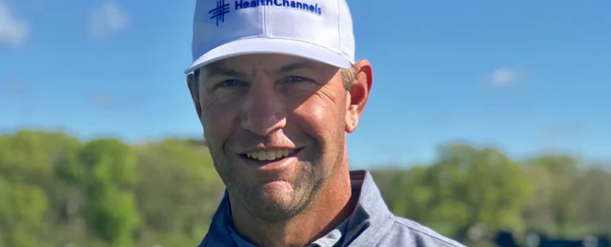 Man wearing a white HealthChannels cap and blue jacket outdoors with trees and blue sky in background