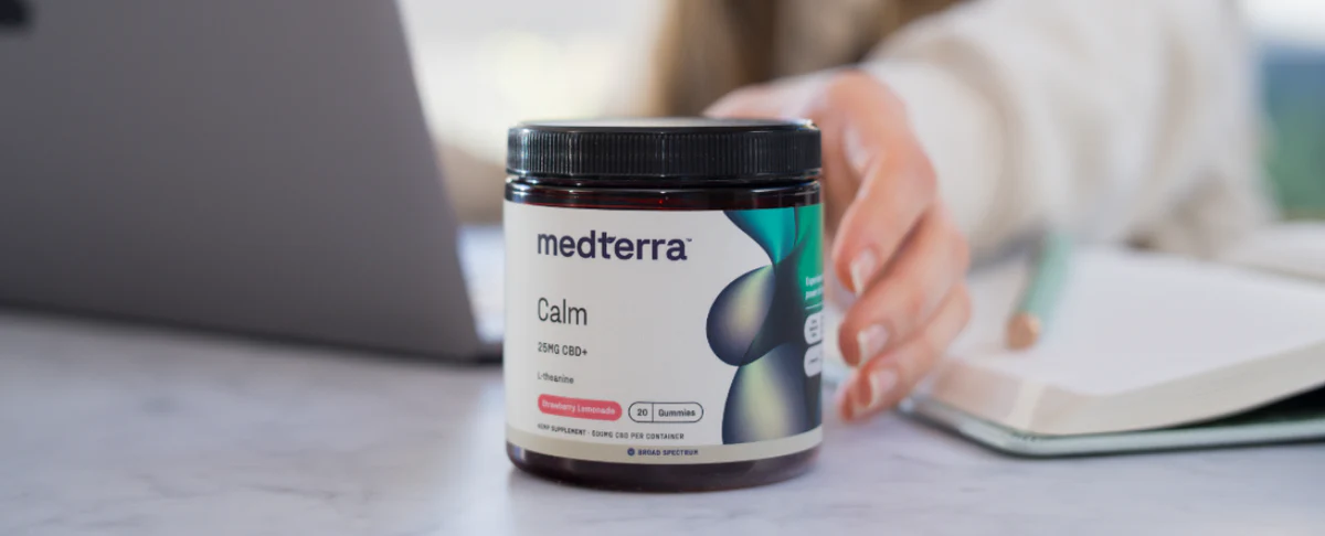 Front view of Medterra Calm gummy jar showing Calm label, 25 mg CBD+ and Strawberry Lemonade flavor with a hand and blurred laptop and notebook in the background