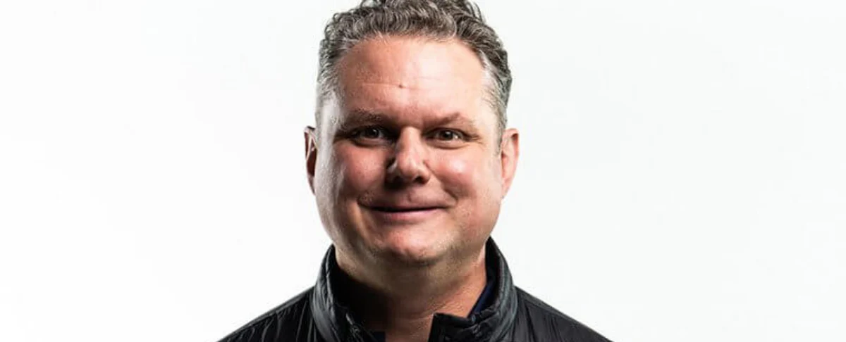 Head-and-shoulders portrait of a smiling man, the company’s operations and supply chain specialist, wearing a black jacket against a white background