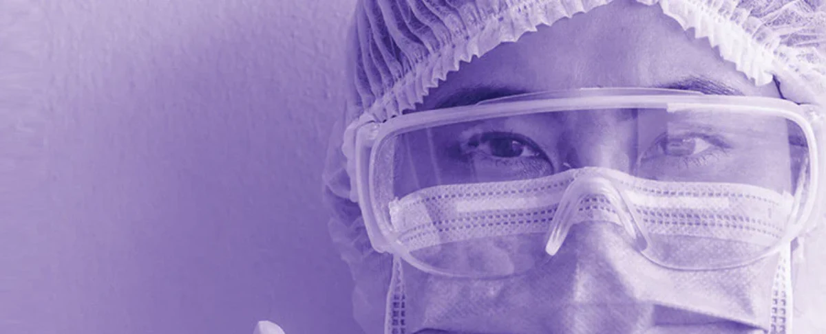 Healthcare worker wearing surgical mask, protective goggles, and hair cover in a purple-toned close-up
