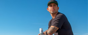 Billy Kemper sitting on the beach holding a CBD product, looking thoughtful against a clear blue sky.