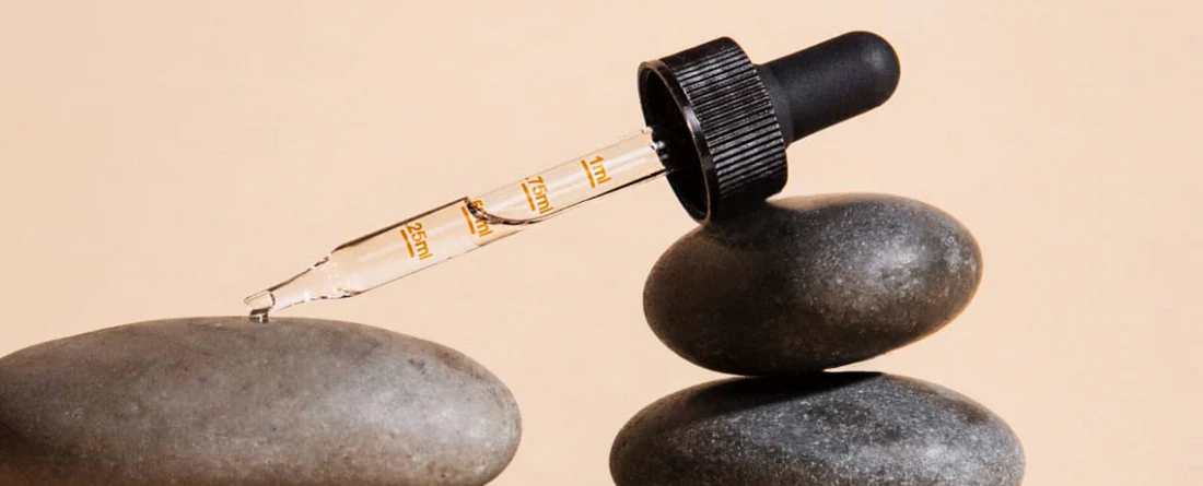 CBD oil dropper releasing a droplet over stacked smooth stones on a beige background, glass pipette showing 0.25–1 ml markings