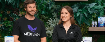 Two smiling people wearing Medterra branded clothing with Medterra CBD products on display in front of a green plant wall background.