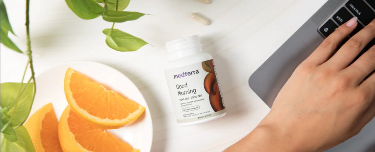 A hand reaching for a bottle of Medterra Good Morning supplements next to orange slices on a plate.