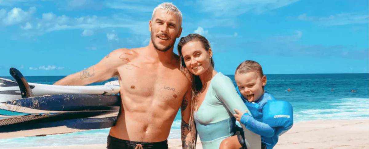 Couple and young child on a sunny beach, man holding surfboards and woman in a light-blue wetsuit holding the child