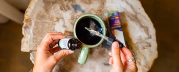 Person holding a dropper over a green mug with CBD oil bottle nearby