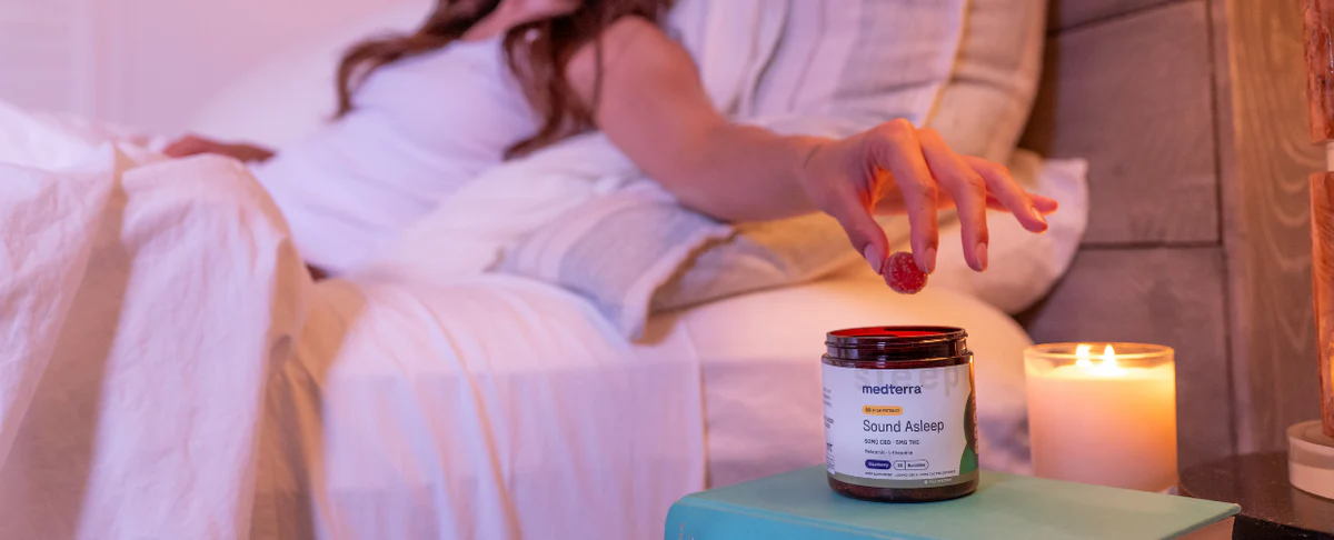 A person reaching for a jar of Sound Asleep CBD Gummies on a bedside table in a cozy bedroom setting.
