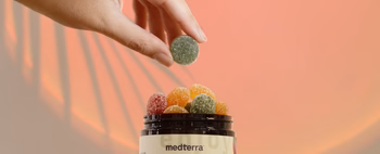 A hand holding a green gummy above a jar filled with colorful CBD gummies.