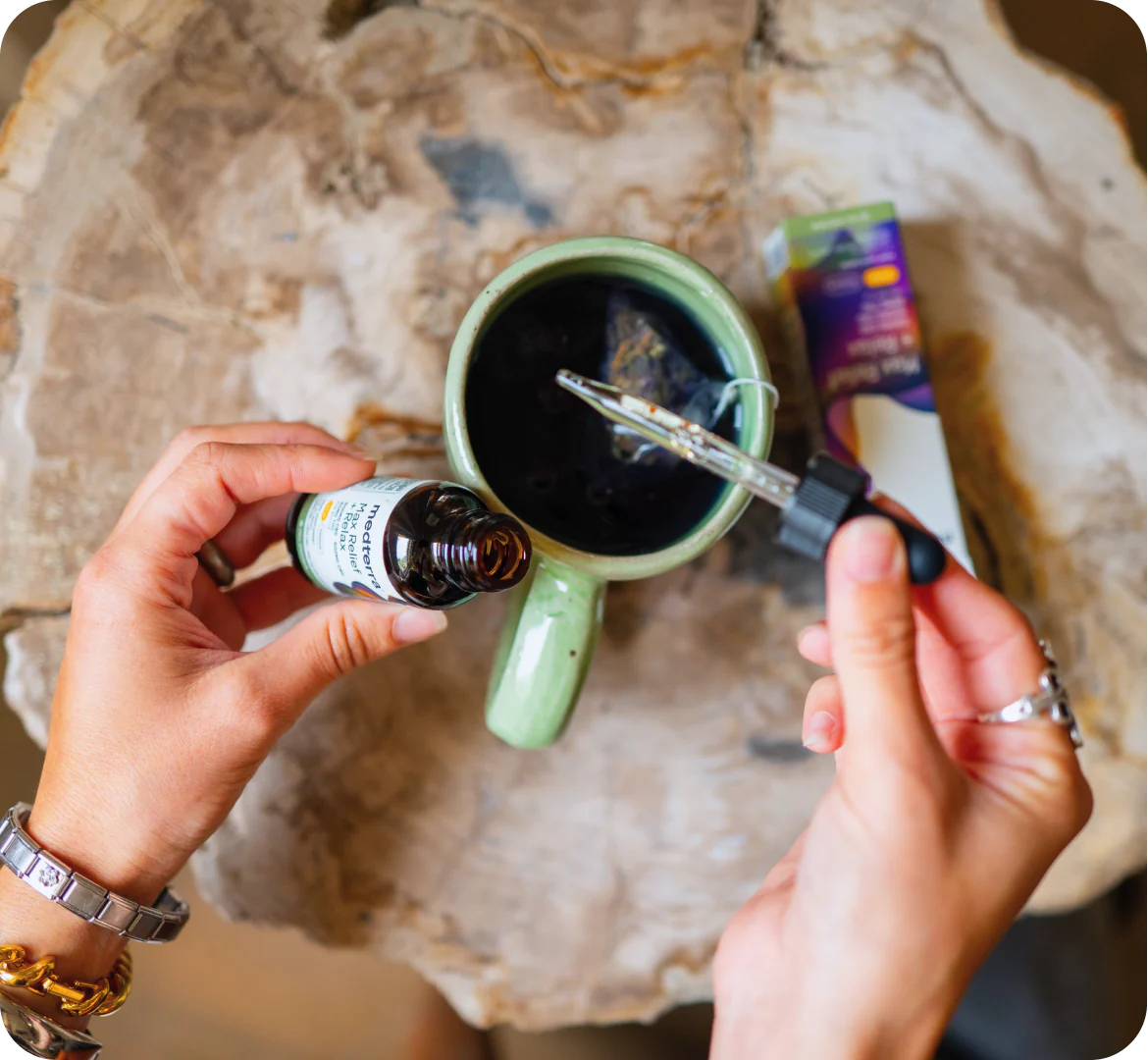 Hands holding Medterra Max Relief tincture bottle and dropper over a green cup with tea on a wooden table
