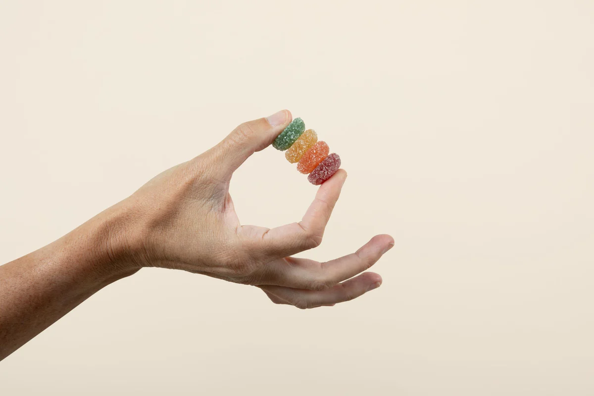Hand holding a stack of four sugar-coated gummy rings (green, yellow, orange, red) against a beige background.