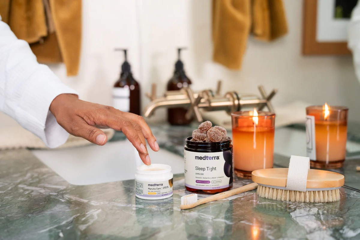 Hand reaching for a Medterra moisturizer jar beside a Medterra Sleep Tight gummies container on a marble bathroom counter with lit candles and a wooden brush