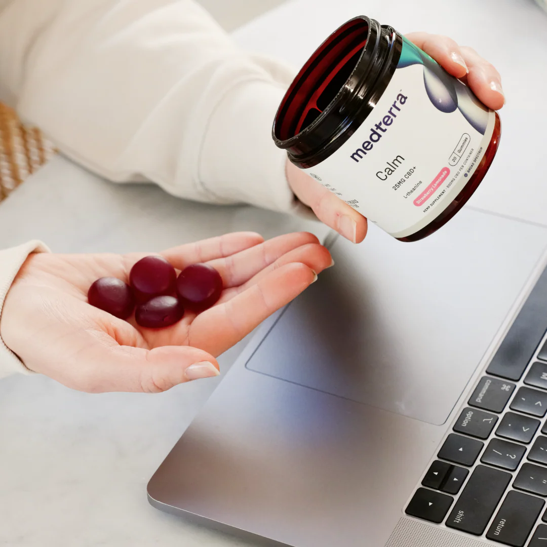 Hand holding four burgundy gummies beside a medterra jar labeled Calm 25MG CBD+ L-Theanine Strawberry Lemonade