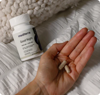 Angled close-up of Medterra Good Night CBD capsules bottle showing the label and two capsules held in a hand on white quilted bedding