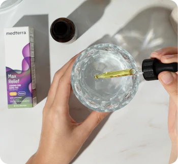 Hands holding a glass while a CBD tincture dropper releases oil; Medterra "Max Relief" box and open bottle at left.