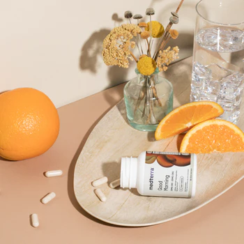 Angled view of Good Morning CBD capsules bottle on its side with spilled capsules on a tray, orange slices and water