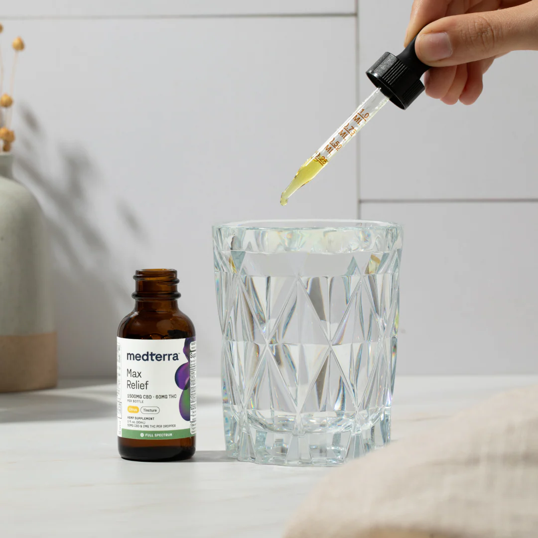 Hand holding a dropper releasing CBD oil into a glass of water next to a small tincture bottle on a countertop