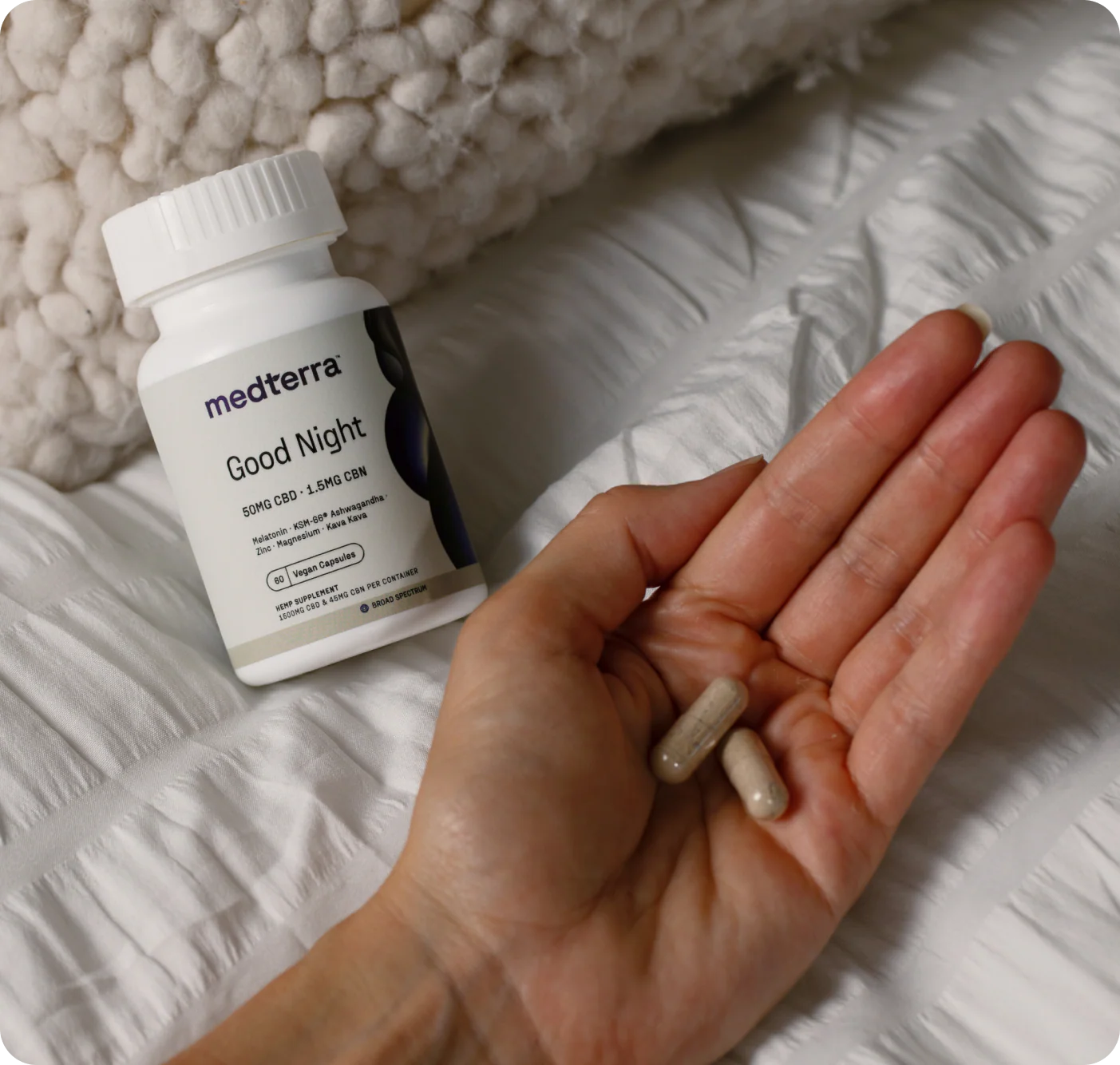 Close-up of Medterra Good Night capsule bottle with a hand holding two capsules on a white quilted bedspread