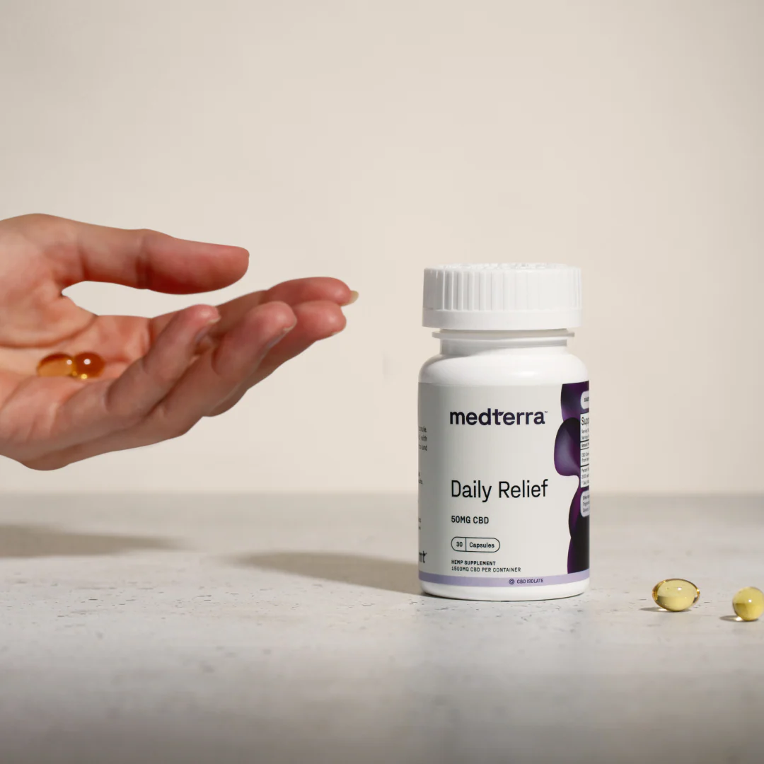 Hand holding two golden softgel capsules above a white bottle of Daily Relief CBD Isolate capsules with a purple label on a light surface