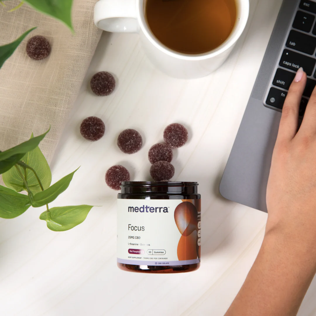 Top-down view of Medterra Focus CBD Gummies jar with sugar-coated red raspberry gummies beside a laptop and tea