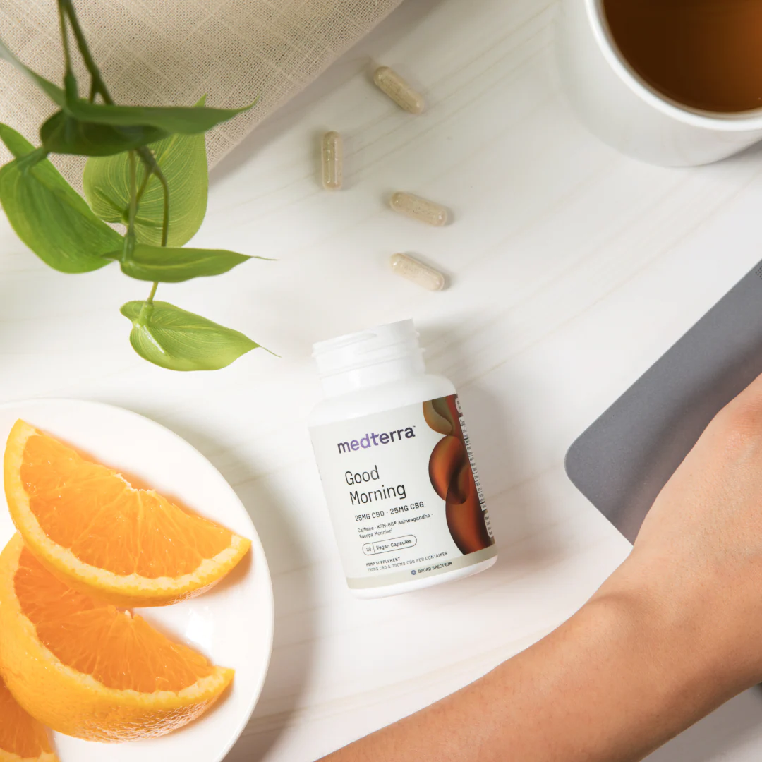 Angled top-down view of Medterra Good Morning bottle with loose capsules, orange slices, tea, and a hand on white tabletop