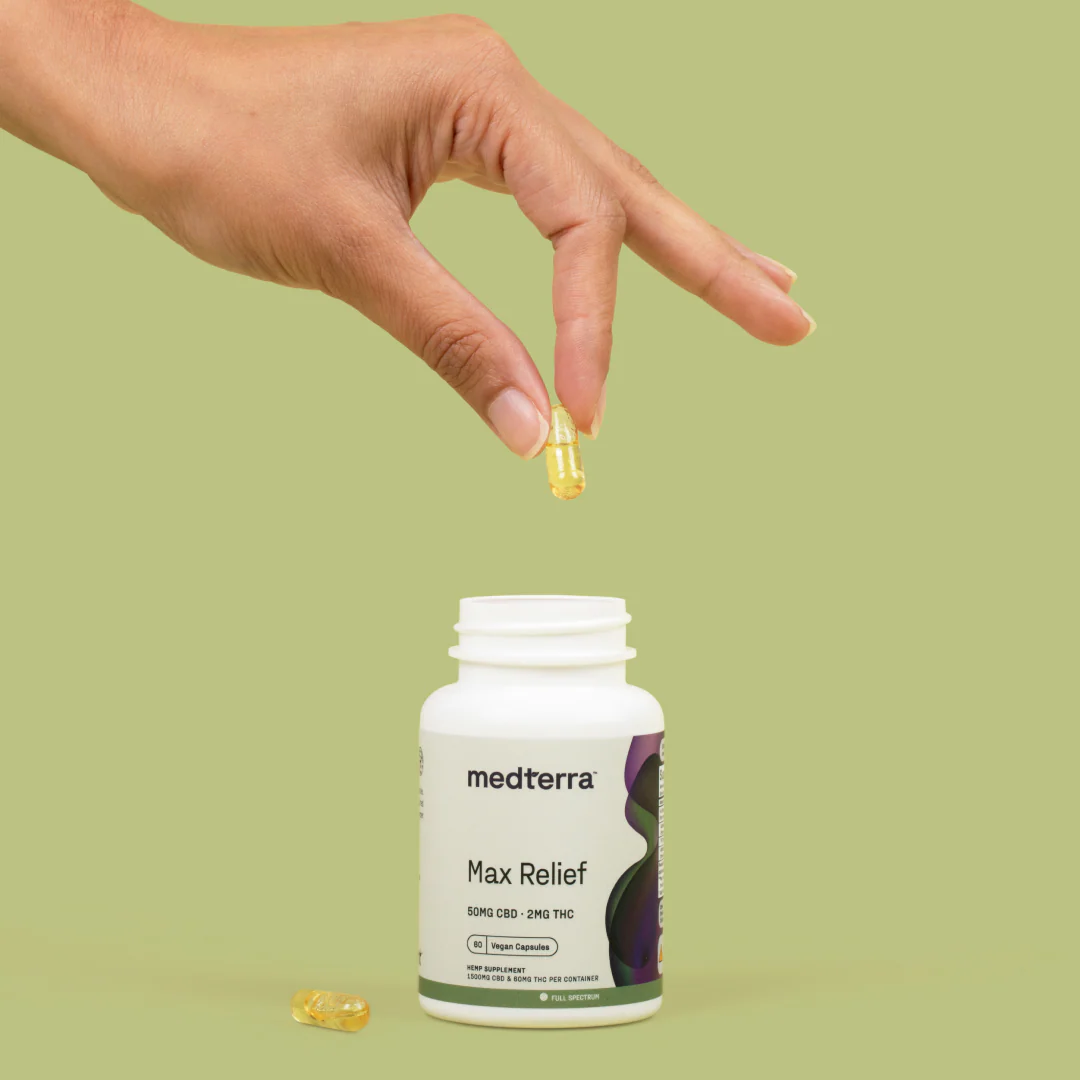 Hand holding Medterra Max Relief full spectrum capsule above open bottle labeled 50 mg CBD, 2 mg THC on green background