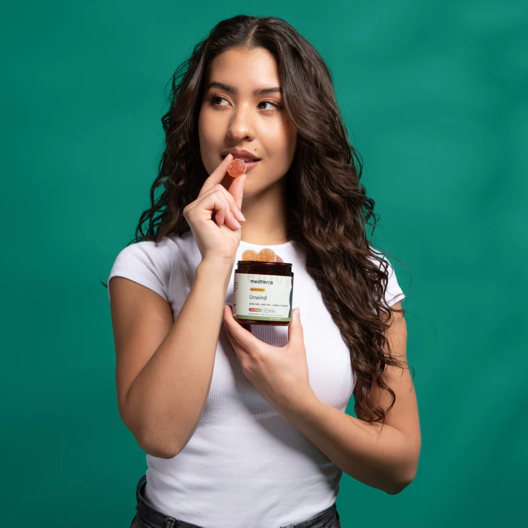 A person with long, wavy hair holds an open container of Medterra Unwind Gummies in one hand and is about to eat one with the other. They are wearing a white t-shirt and standing against a solid green background.