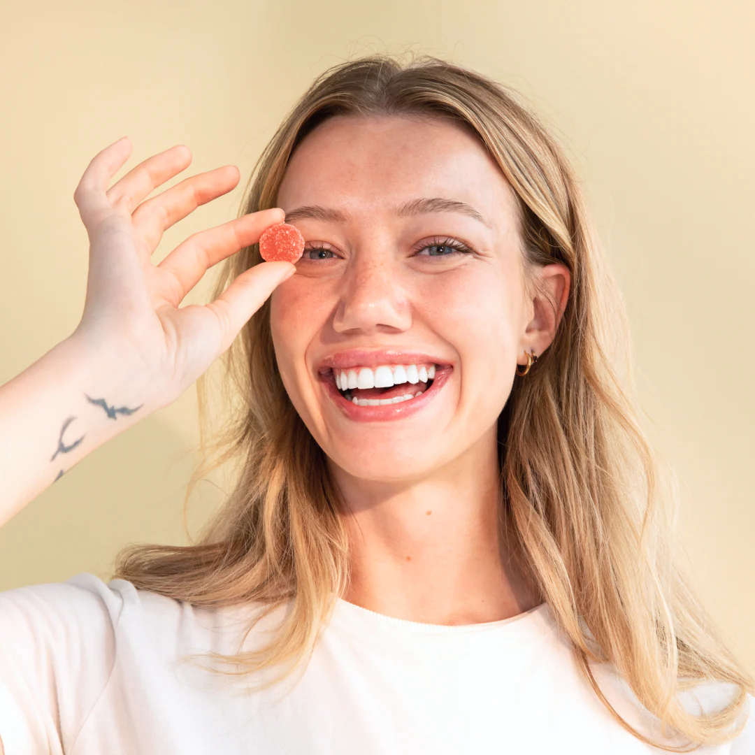 Smiling woman holding a red Daily Delight CBD gummy over her right eye against a muted yellow-beige background