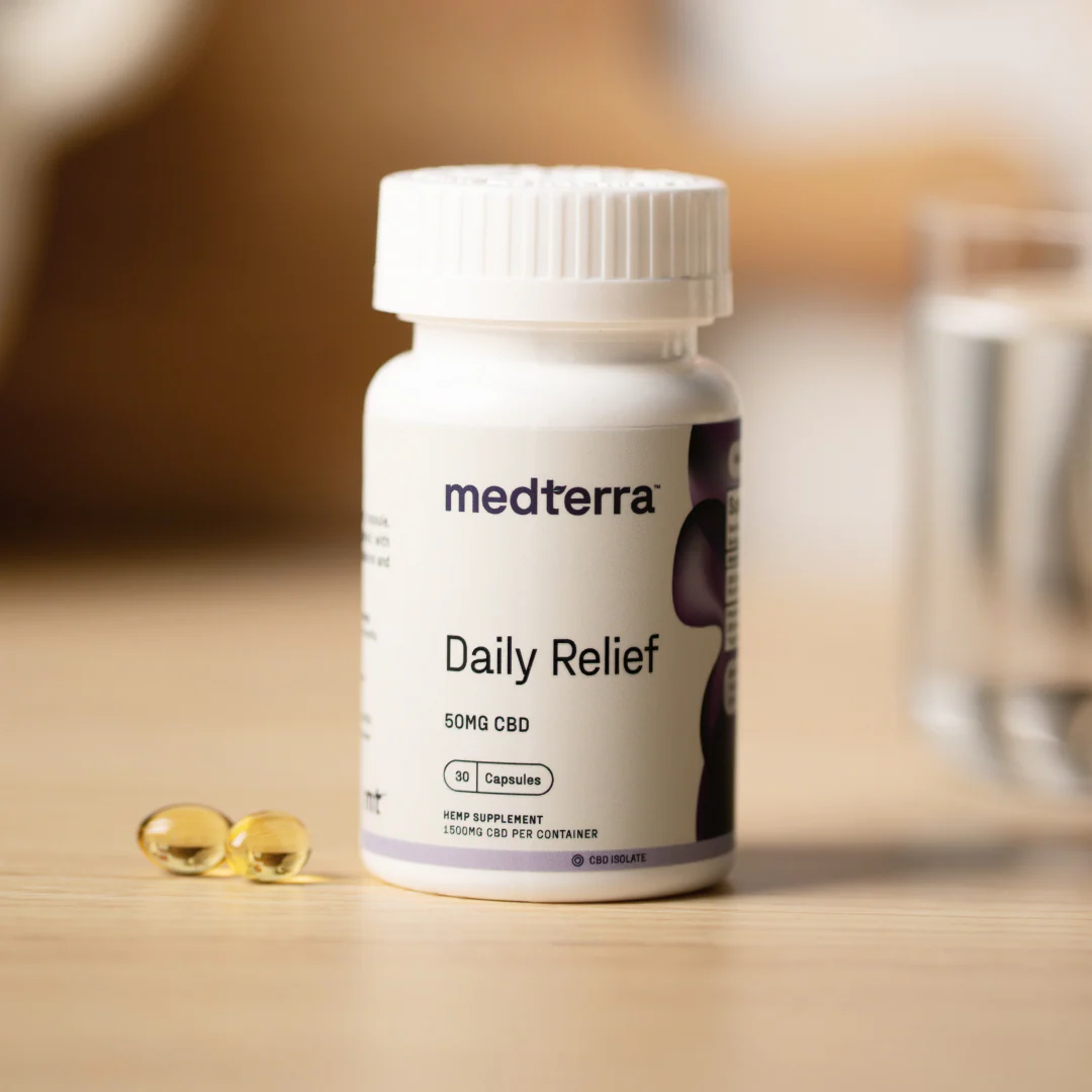 Front view of Daily Relief 50MG CBD isolate bottle with two golden capsules on a wooden table and blurred glass of water