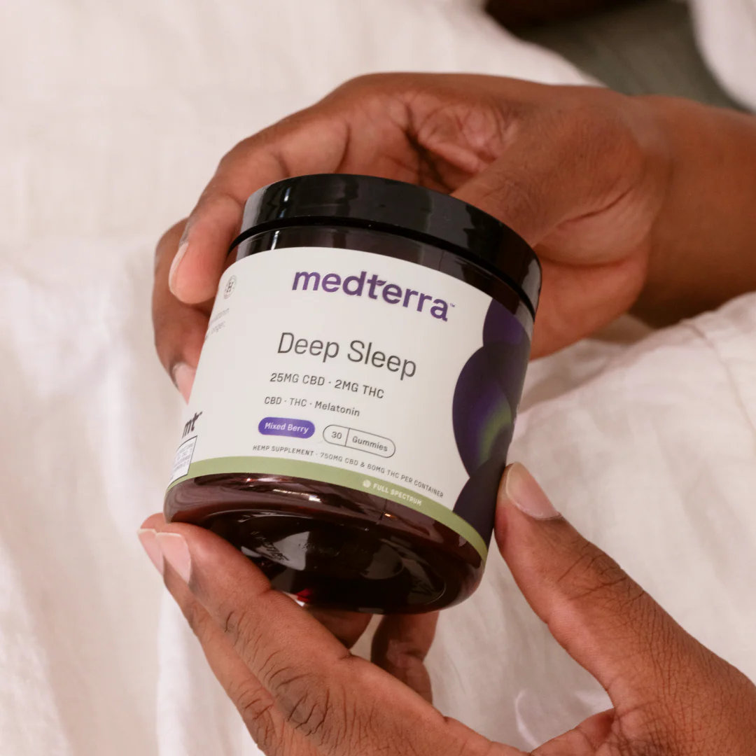 Angled view of Medterra Deep Sleep gummies jar showing label 25 mg CBD, 2 mg THC and mixed berry flavor, held in hands on white bedding