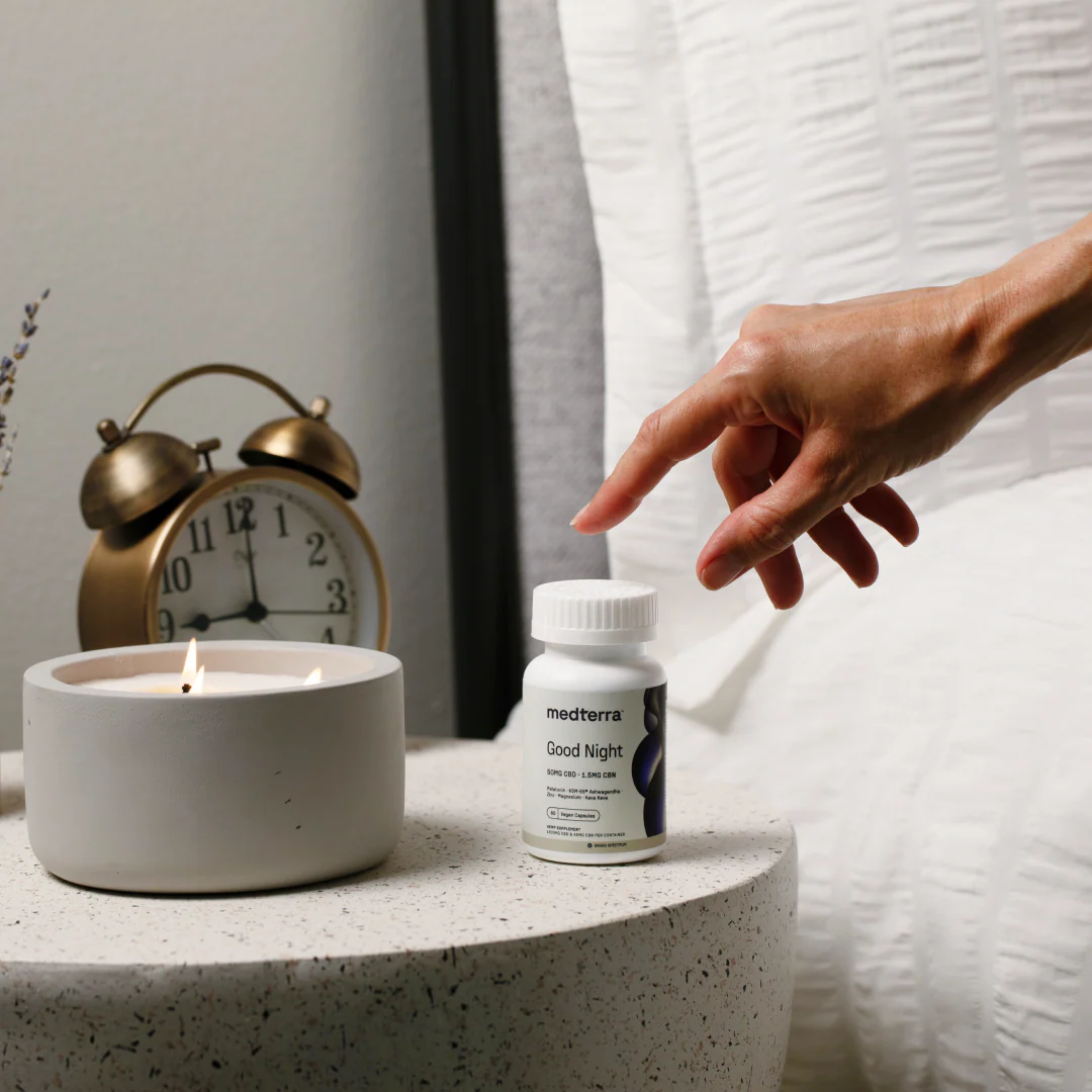 Hand reaching for Medterra Good Night capsules on a bedside table with a lit candle and an alarm clock