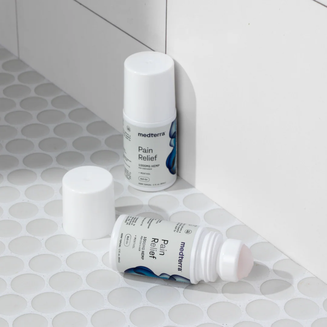 Two Medterra Pain Relief Roll-On bottles, each containing 1000mg of CBD, are displayed against a white tiled surface. One stands upright with its cap on, while the other lies on its side with the cap off, exposing the roll-on applicator crafted for joint pain relief and enduring comfort.