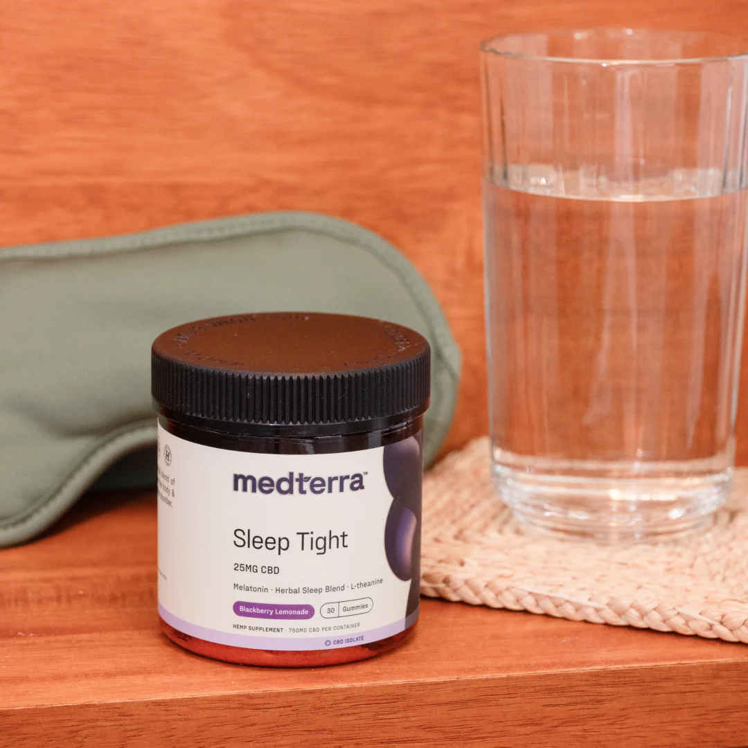 Jar of Medterra Sleep Tight CBD gummies, 25 mg, on a wooden surface with a glass of water and a green sleep mask