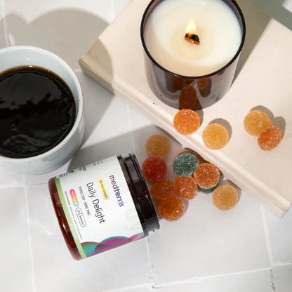 Spilled jar of Daily Delight CBD Gummies beside a cup of black coffee and a lit candle on white tile