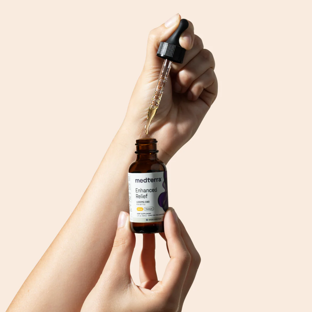 Close-up of Medterra Enhanced Relief oil bottle held in hands, dropper dispensing oil against pale peach background