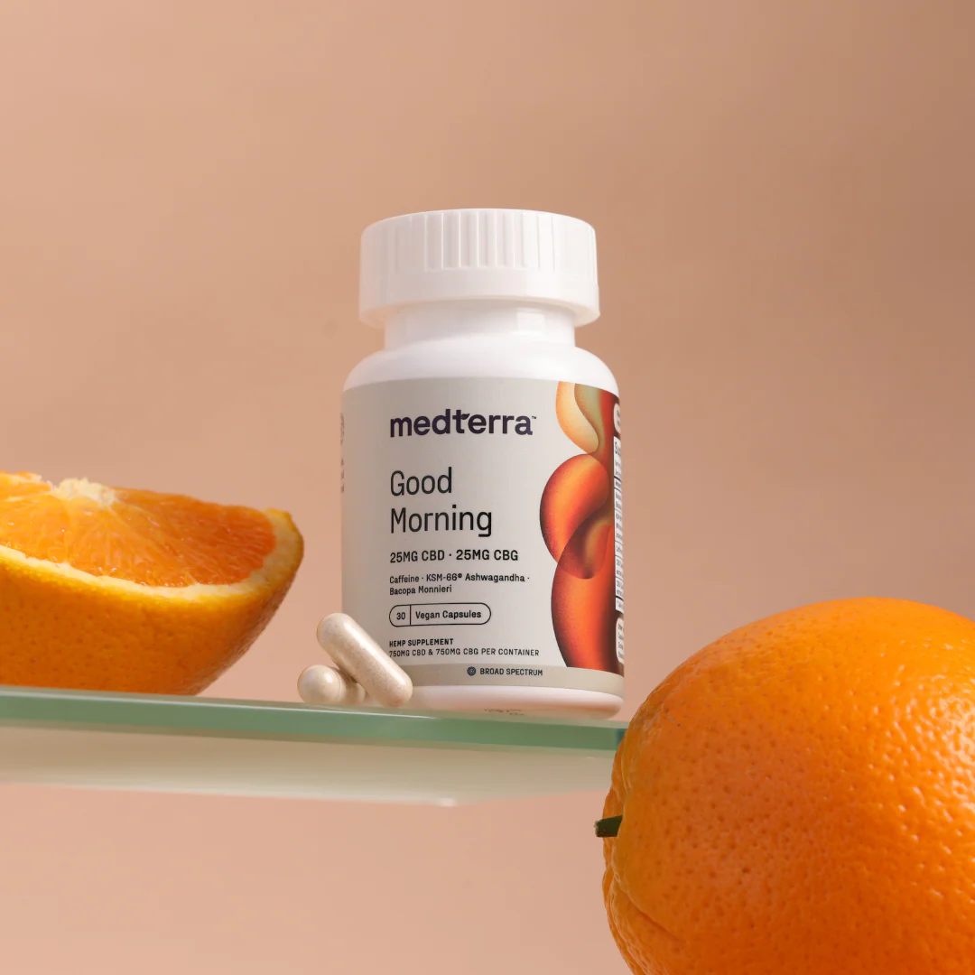Front view of Medterra Good Morning bottle with two vegan capsules on glass shelf, orange and orange slice, peach background