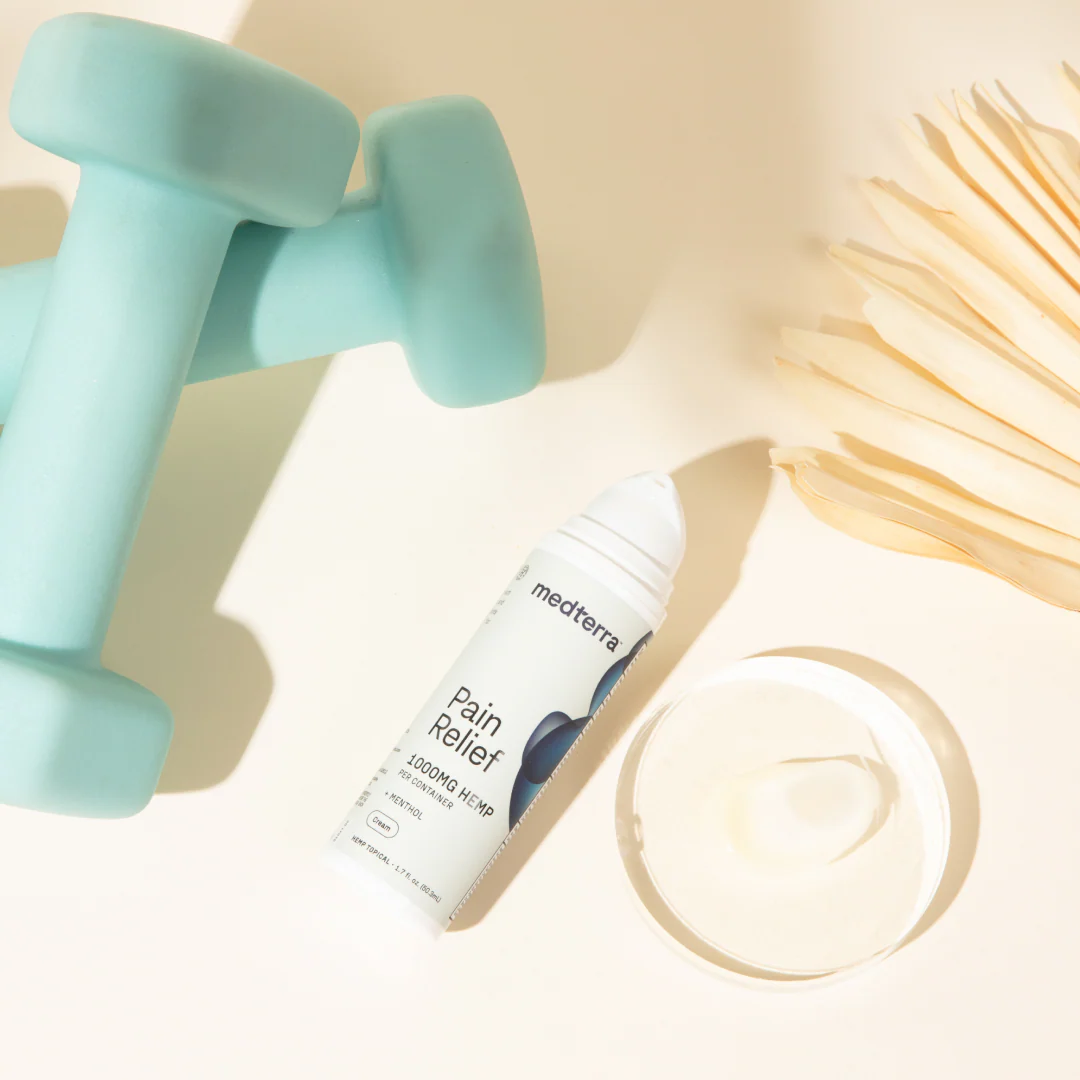 Angled overhead of Pain Relief Cream tube and open jar with cream next to mint dumbbells and palm leaf on pale background