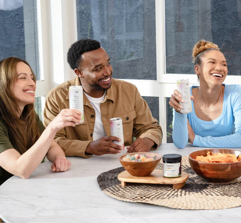 Three friends laughing and sharing snacks and canned CBD drinks at a bright kitchen table with a jar of CBD products