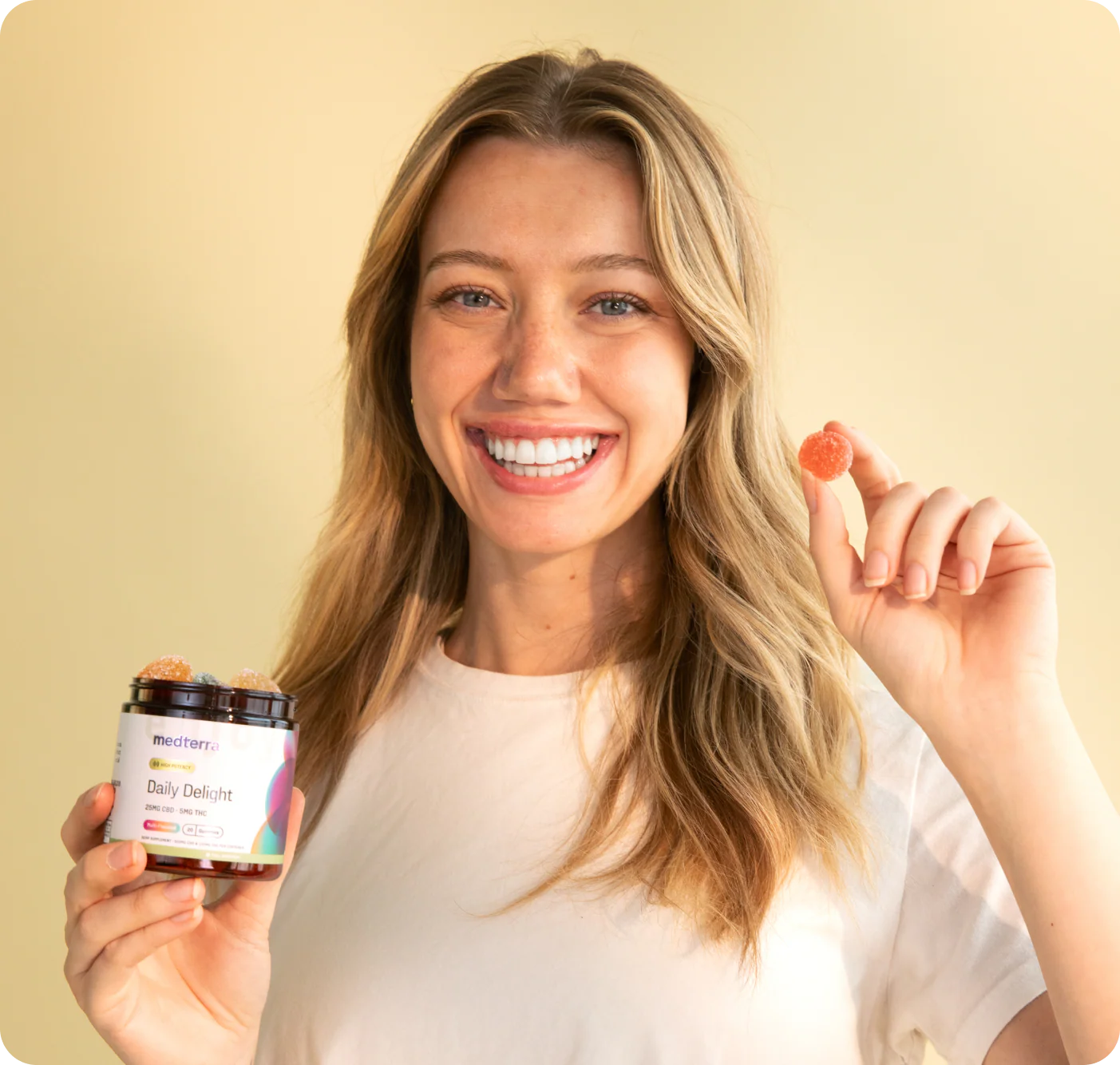 Smiling woman holding a jar of Daily Delight gummies and displaying one orange gummy against a pale beige background