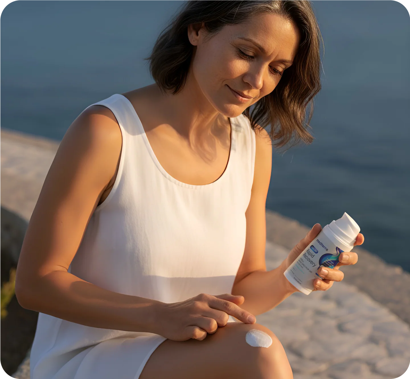 Woman by the water applying white cream to her knee while holding a topical lotion bottle.
