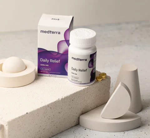 Three quarter view of Daily Relief CBD bottle and box with three yellow softgel capsules on beige geometric props