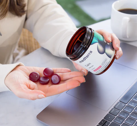 Close-up of jar of Medterra Calm Ultra Broad Spectrum CBD gummies showing red gummies in a hand beside a laptop and coffee cup