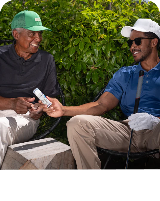 Two men outdoors exchanging a small white bottle, one wearing a green cap and the other a white cap and sunglasses, with greenery behind them.