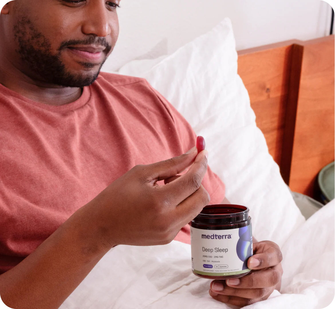 Man holding a red sleep gummy and an open jar of Medterra Deep Sleep gummies while sitting in bed with white bedding