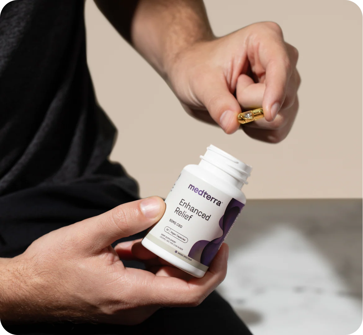 Close-up of hands holding an open Medterra Enhanced Relief bottle and a gold capsule above it on a blurred background