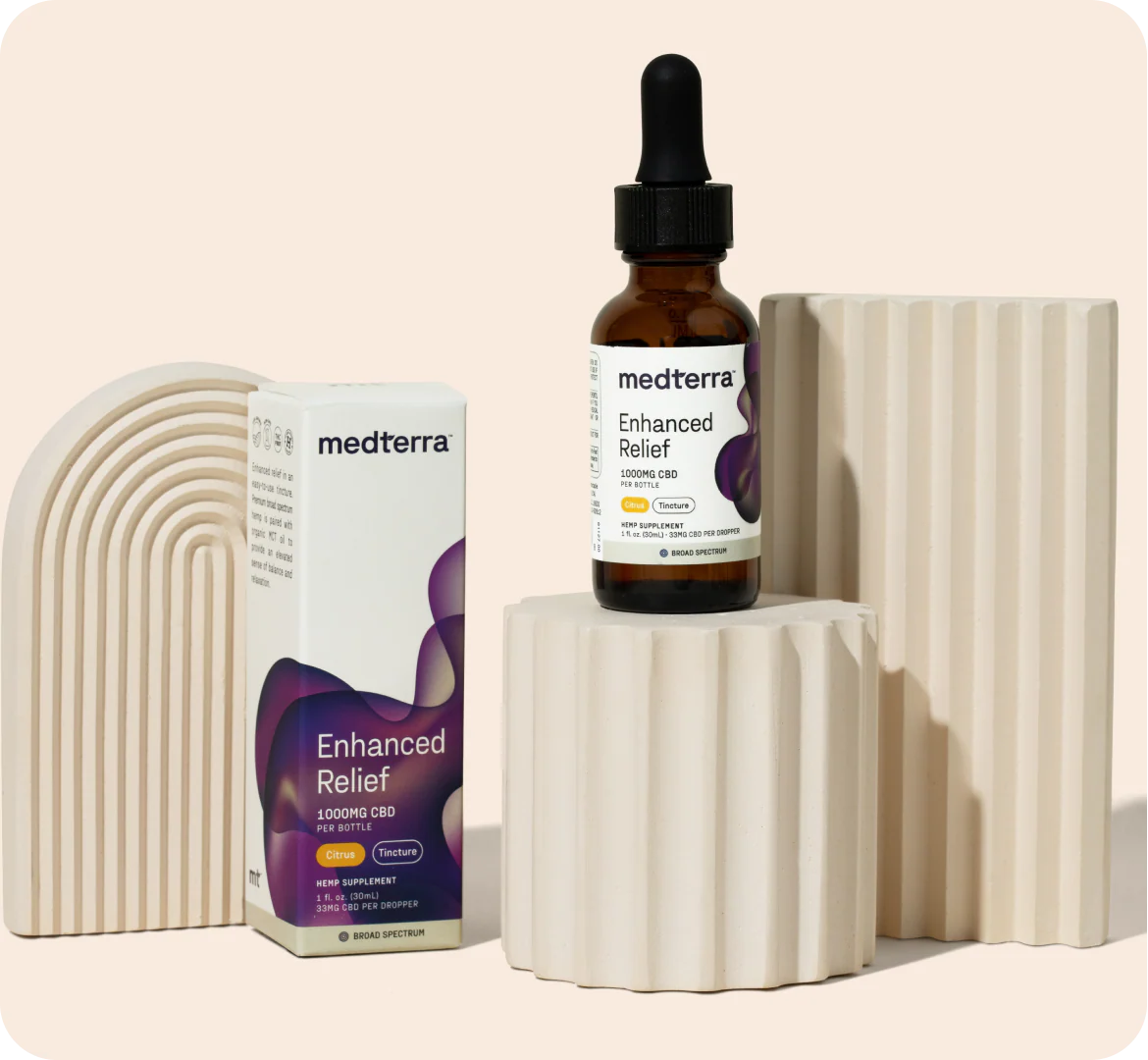 Front view of Enhanced Relief 1000mg CBD tincture bottle and box on beige ribbed pedestals showing dropper and purple label