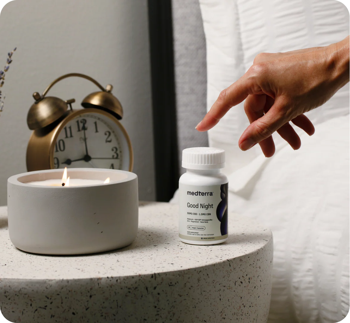Close-up of Medterra Good Night capsule bottle showing the Good Night label on a bedside table with lit candle, brass alarm clock, and hand reaching