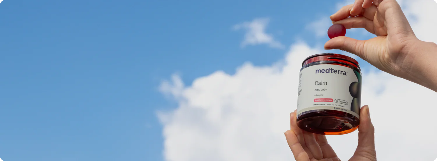 Close-up of Medterra Calm gummies jar held by hands, open with one pink gummy above it against blue sky