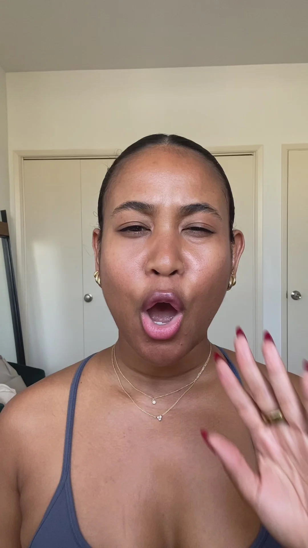 A person with pulled-back hair making an expressive face with mouth open and raising their hand near their face, wearing gold earrings and layered necklaces.