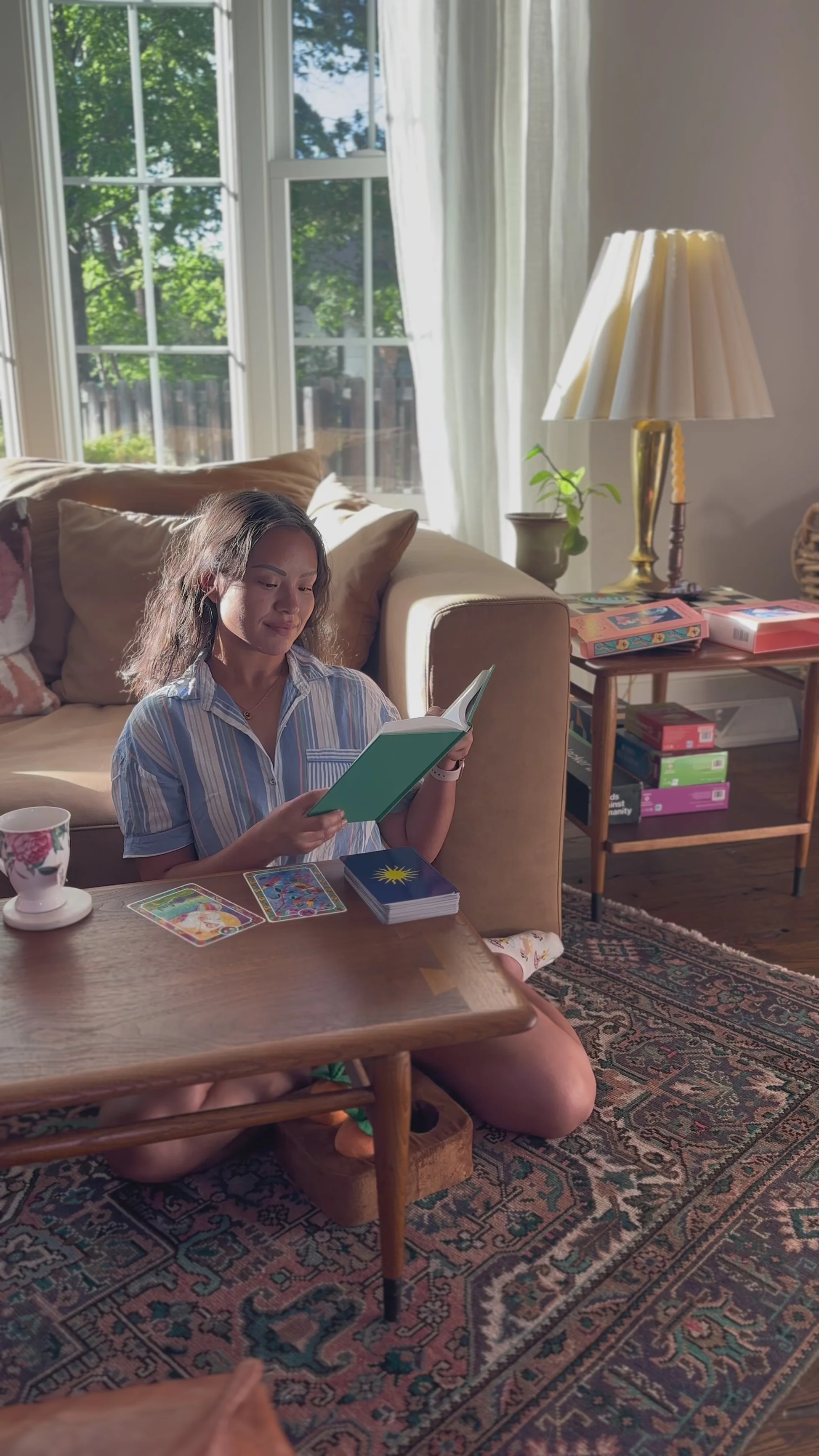 Woman sitting on the floor reading a book with tarot cards spread on a wooden table in a sunlit living room.